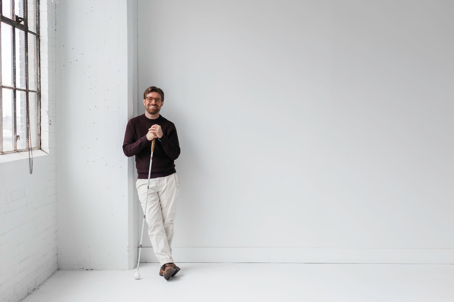 Charlie Kramer leaning against a bright studio wall by a tall window, smiling warmly while resting both hands on a white cane.