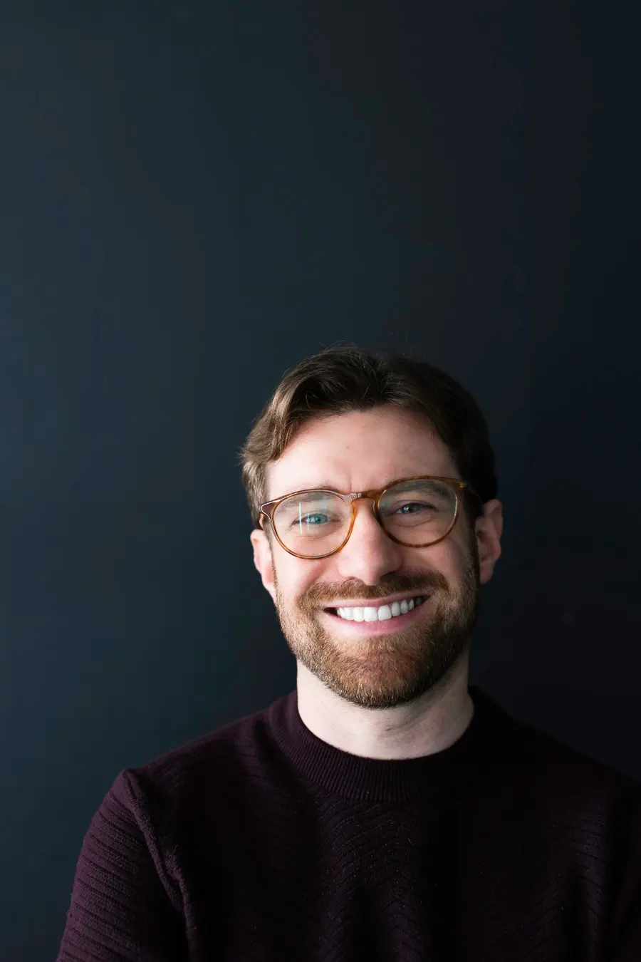 Portrait of Charlie Kramer smiling warmly, wearing tortoiseshell glasses and a burgundy sweater against a deep navy background.