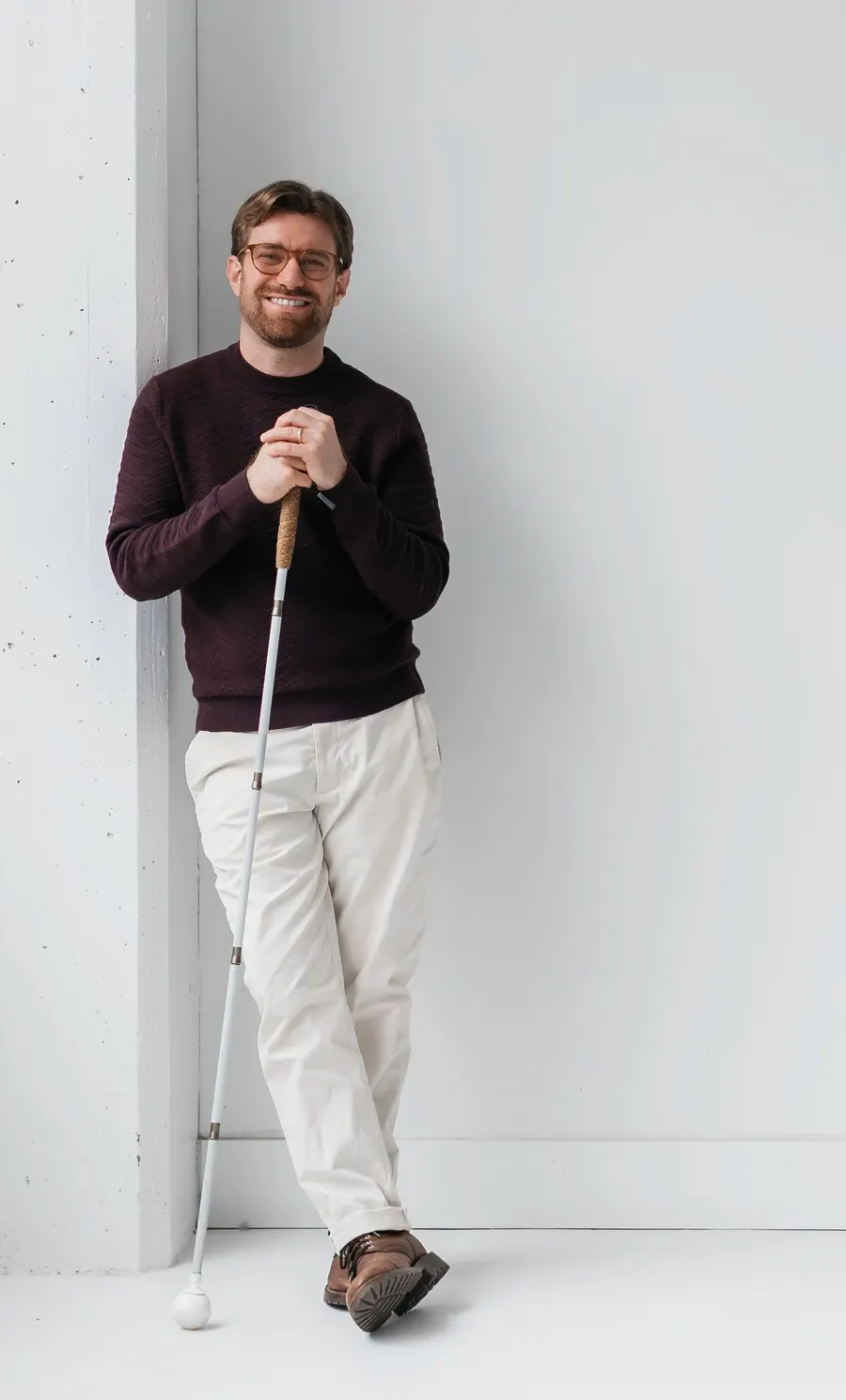 Charlie Kramer leaning against a bright studio wall by a tall window, smiling warmly while resting both hands on a white cane.